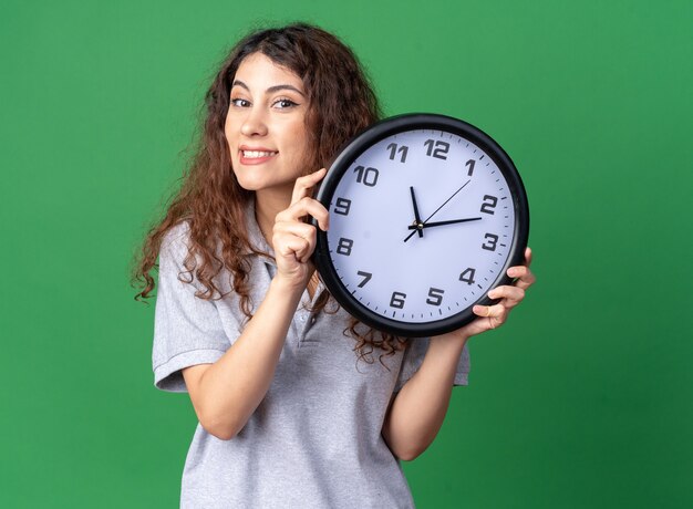 Smiling young pretty woman holding clock looking at front isolated on green wall