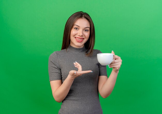 Smiling young pretty woman holding and pointing at cup isolated on green background with copy space