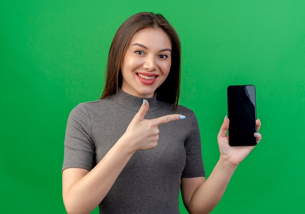 Smiling young pretty woman holding and pointing at mobile phone isolated on green background