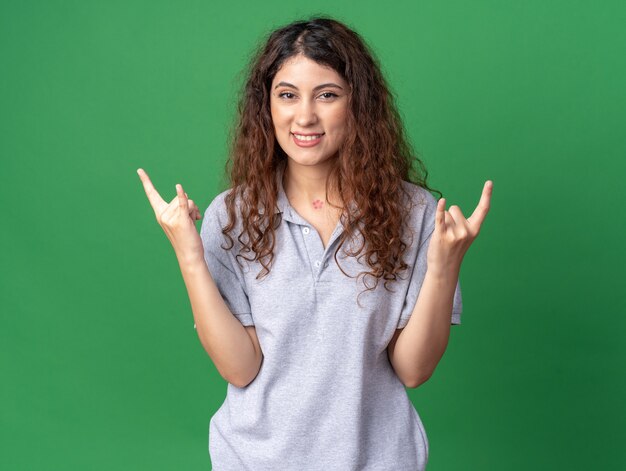 Smiling young pretty woman looking at front doing rock sign isolated on green wall