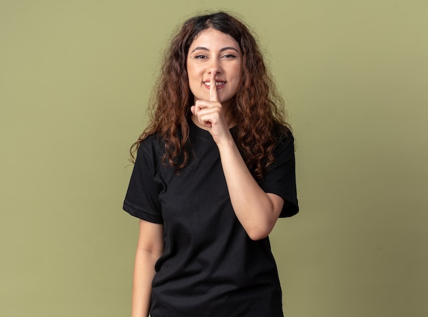 Smiling young pretty woman looking at front doing silence gesture isolated on olive green wall with copy space