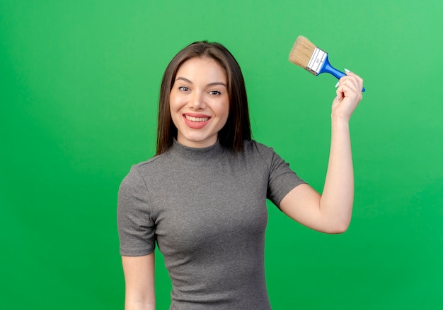Smiling young pretty woman raising up paint brush isolated on green background with copy space