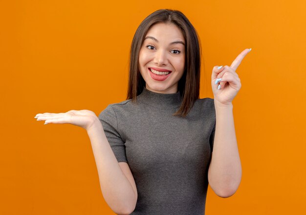 空の手を示し、オレンジ色の背景で隔離の側を指して笑顔の若いきれいな女性