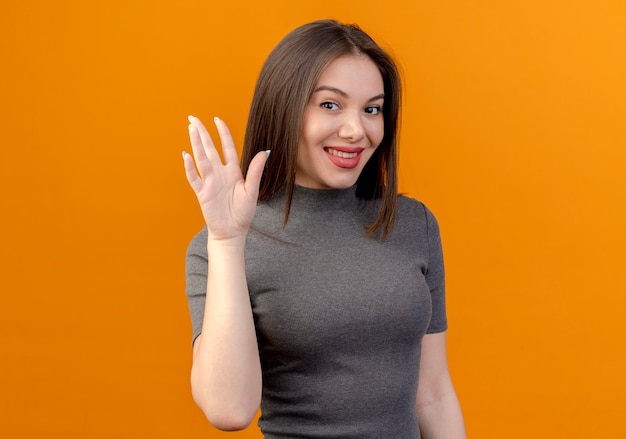 Smiling young pretty woman waving at camera isolated on orange background with copy space