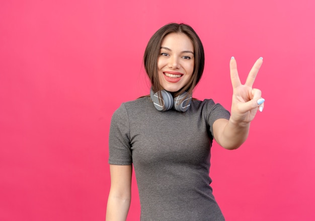 Sorridente giovane donna graziosa che indossa le cuffie sul collo facendo segno di pace isolato su sfondo rosa con spazio di copia