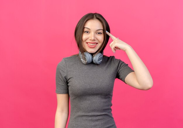 Smiling young pretty woman wearing headphones on neck putting finger on temple isolated on pink background with copy space