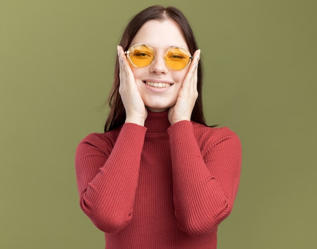 올리브 녹색 벽에 고립 된 전면을보고 얼굴에 손을 유지 선글라스를 착용 웃는 젊은 예쁜 여자