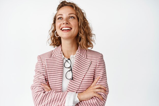 Smiling young professional, businesswoman looking at upper left corner with hopeful face expression, standing on white.