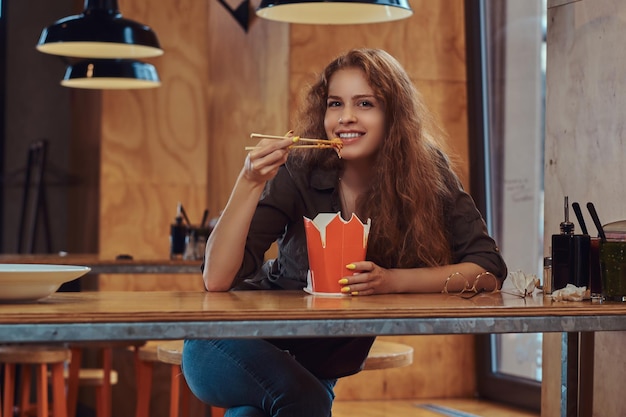 Sorridente giovane donna rossa che indossa abiti casual che mangiano noodles piccanti in un ristorante asiatico.