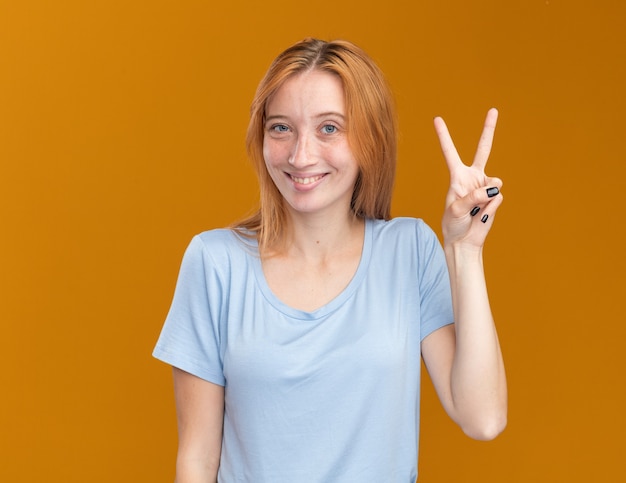 Smiling young redhead ginger girl with freckles gesturing victory sign isolated on orange wall with copy space