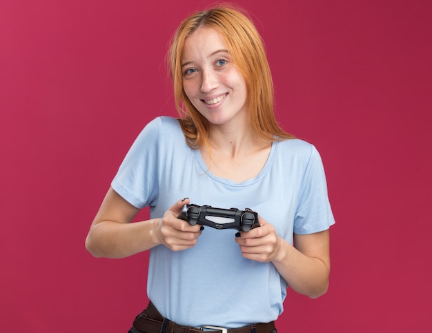 Smiling young redhead ginger girl with freckles holding game controller on pink