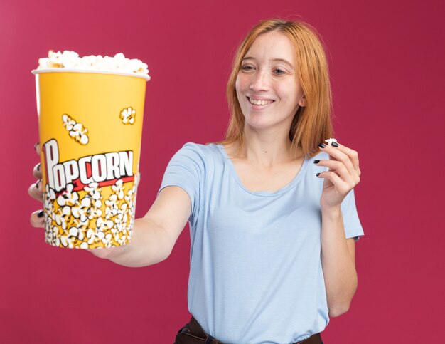 Smiling young redhead ginger girl with freckles holding and looking at popcorn bucket