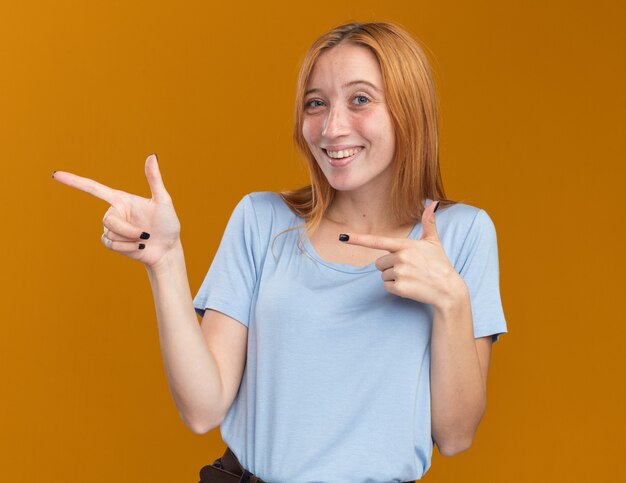Smiling young redhead ginger girl with freckles pointing at side isolated on orange wall with copy space