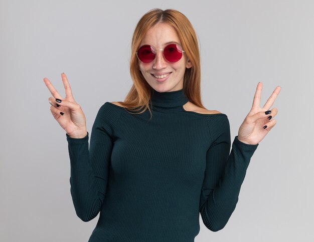 Smiling young redhead ginger girl with freckles in sun glasses gesturing victory sign on white