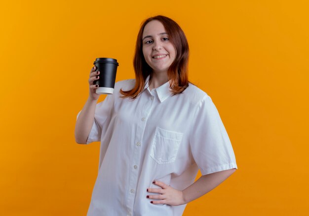 Smiling young redhead girl holding cup of coffee and putting hand on hip isolated on yellow background