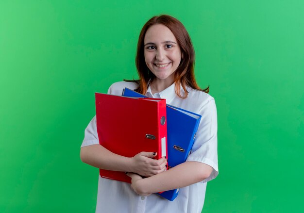 Smiling young redhead girl holding folders isolated on green