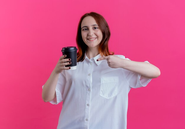 Smiling young redhead girl holding and points at cup of coffee isolated on pink