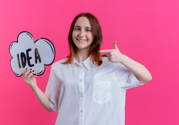 Smiling young redhead girl holding and points at idea bubble isolated on pink