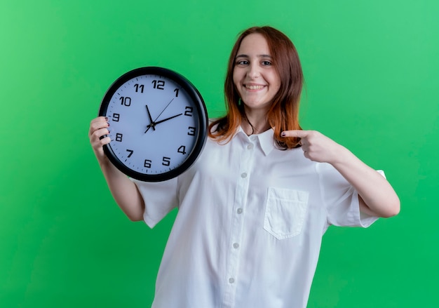 Smiling young redhead girl holding and points at wall clock isolated on green