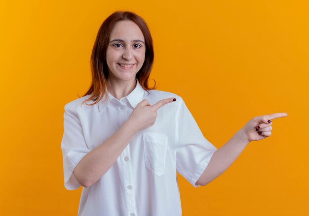 Smiling young redhead girl points at side  isolated on yellow with copy space