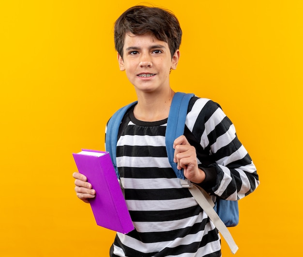 smiling young school boy wearing backpack holding book isolated on orange wall