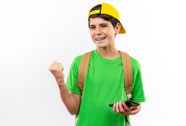 Smiling young school boy wearing backpack with cap holding phone showing yes gesture isolated on white wall
