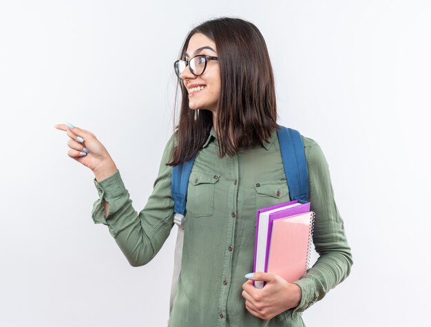 Smiling young school woman wearing glasses with backpack holding books points at side 