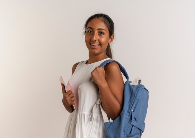 Smiling young schoolgirl wearing back bag holding notebook on white