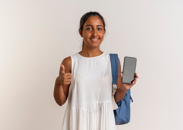Улыбающаяся молодая школьница в сумке на спине, держа телефон большим пальцем вверх, изолирована на белом