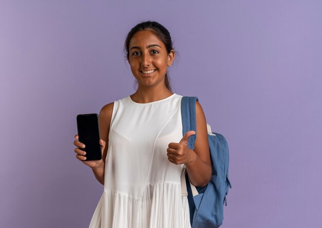 Улыбающаяся молодая школьница в сумке на спине держит телефон большим пальцем на фиолетовом