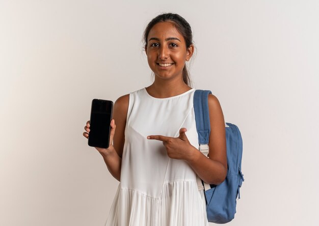 Smiling young schoolgirl wearing back bag holding and points at phone on white