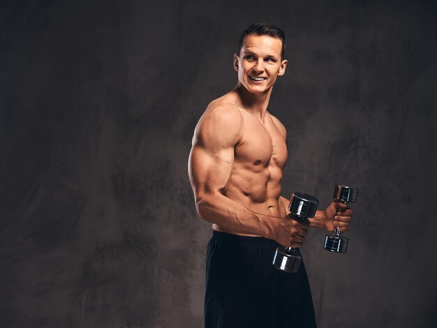 Smiling young shirtless bodybuilder with muscular body doing exercise with dumbbells on a dark background.