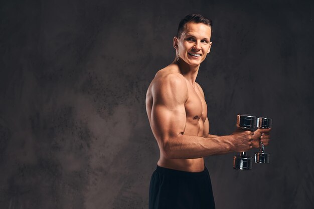 Smiling young shirtless bodybuilder with muscular body doing exercise with dumbbells on a dark background.