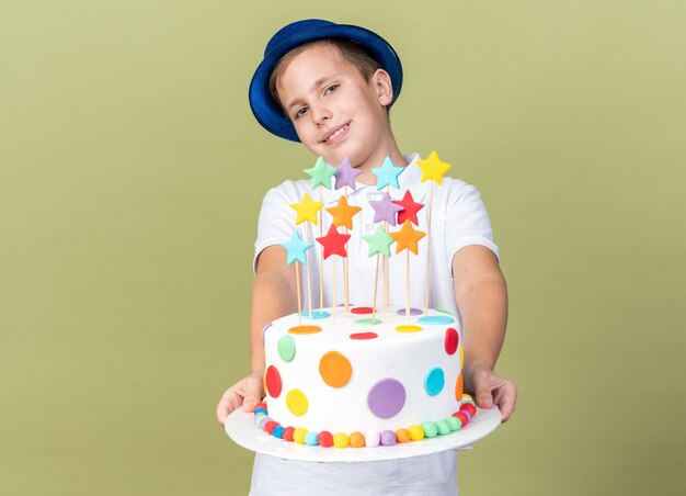 smiling young slavic boy with blue party hat holding birthday cake isolated on olive green wall with copy space