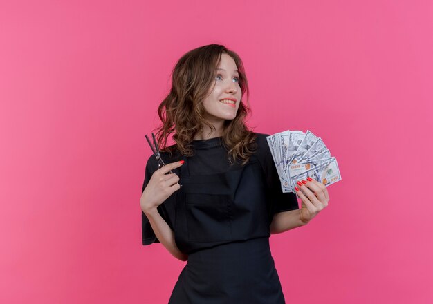 Бесплатное фото Улыбающаяся молодая славянская женщина-парикмахер в униформе смотрит на кусающую губу, держащую деньги и ножницы, изолированные на розовом фоне с копией пространства