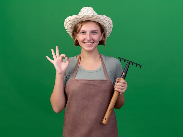 Sorridente giovane giardiniere femminile slavo che indossa il cappello di giardinaggio gesticolando segno ok e tenendo il rastrello sul verde