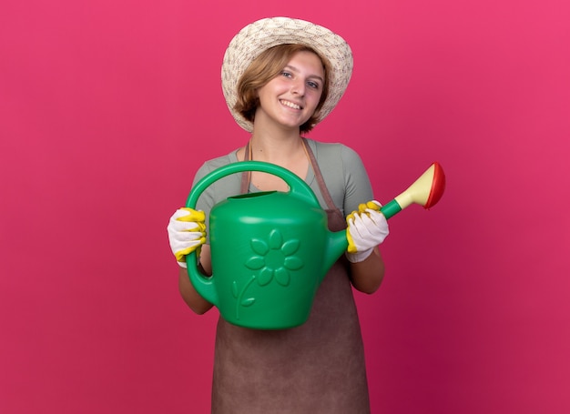 Smiling young slavic female gardener wearing gardening hat and gloves holding watering can isolated on pink wall with copy space