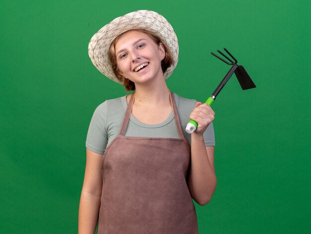 Улыбающаяся молодая славянская женщина-садовник в садовой шляпе держит грабли на зеленом