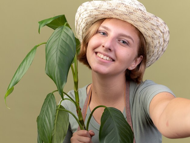 Smiling young slavic female gardener wearing gardening hat holding plant and pretending to hold camera taking selfie