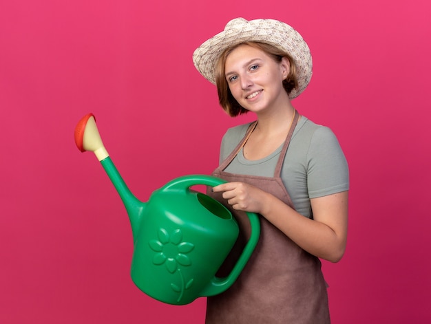 Sorridente giovane giardiniere femminile slavo che indossa cappello da giardinaggio tenendo annaffiatoio sul rosa