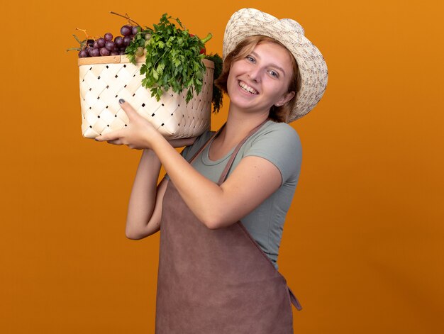 Улыбающаяся молодая славянская женщина-садовник в садовой шляпе держит корзину с овощами, изолированную на оранжевой стене с копией пространства