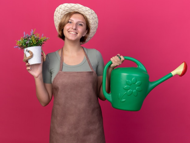 Smiling young slavic female gardener wearing gardening hat holds watering can and flowers in flowerpot on pink