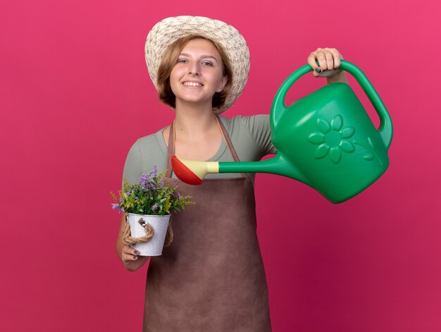 Smiling young slavic female gardener wearing gardening hat watering flowers in flowerpot with watering can isolated on pink wall with copy space