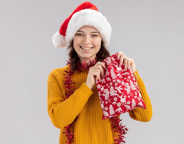 サンタの帽子とコピースペースで白い壁に分離されたクリスマスギフトバッグを保持している首の周りの花輪と笑顔の若いスラブの女の子