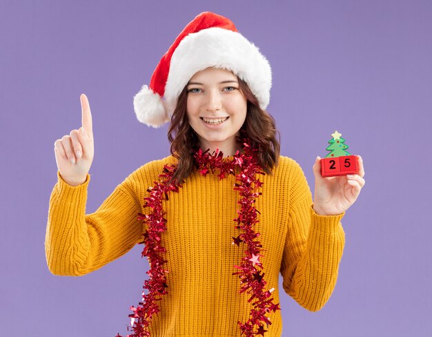 Smiling young slavic girl with santa hat and with garland around neck holding christmas tree ornament and pointing up isolated on purple background with copy space