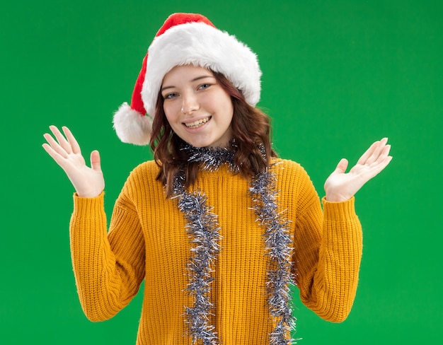 Smiling young slavic girl with santa hat and with garland around neck holding hands open isolated on green wall with copy space