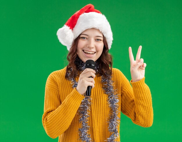 Free photo smiling young slavic girl with santa hat and with garland around neck holding mic and gesturing victory sign