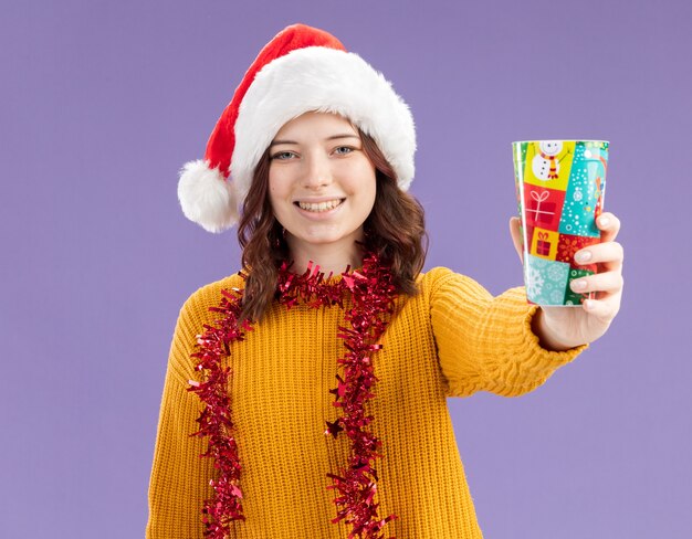 Smiling young slavic girl with santa hat and with garland around neck holding paper cup isolated on purple background with copy space