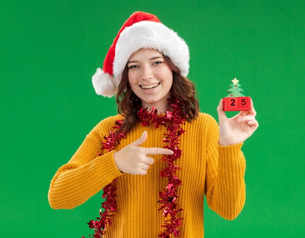 Smiling young slavic girl with santa hat and with garland around neck holding and pointing at christmas tree ornament isolated on green wall with copy space