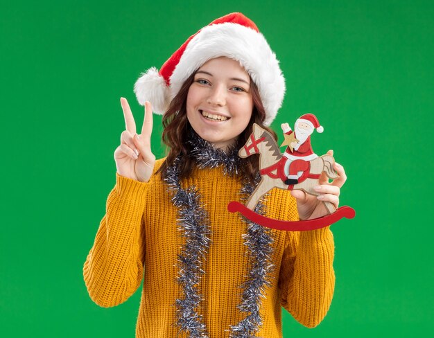 Smiling young slavic girl with santa hat and with garland around neck holding santa on rocking horse decoration and gesturing victory sign 